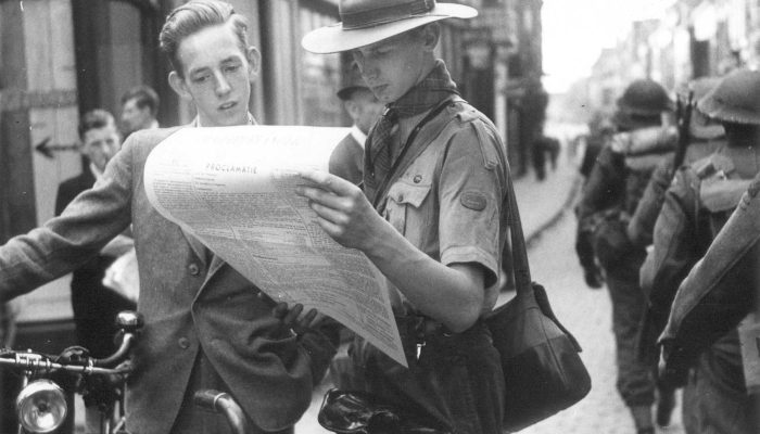 Uitreiking van de eerste Gelderlander krant aan burgers van Nijmegen na de bevrijding van Nijmegen_ aan de rechterkant passeert een groep Britse soldaten. 1944